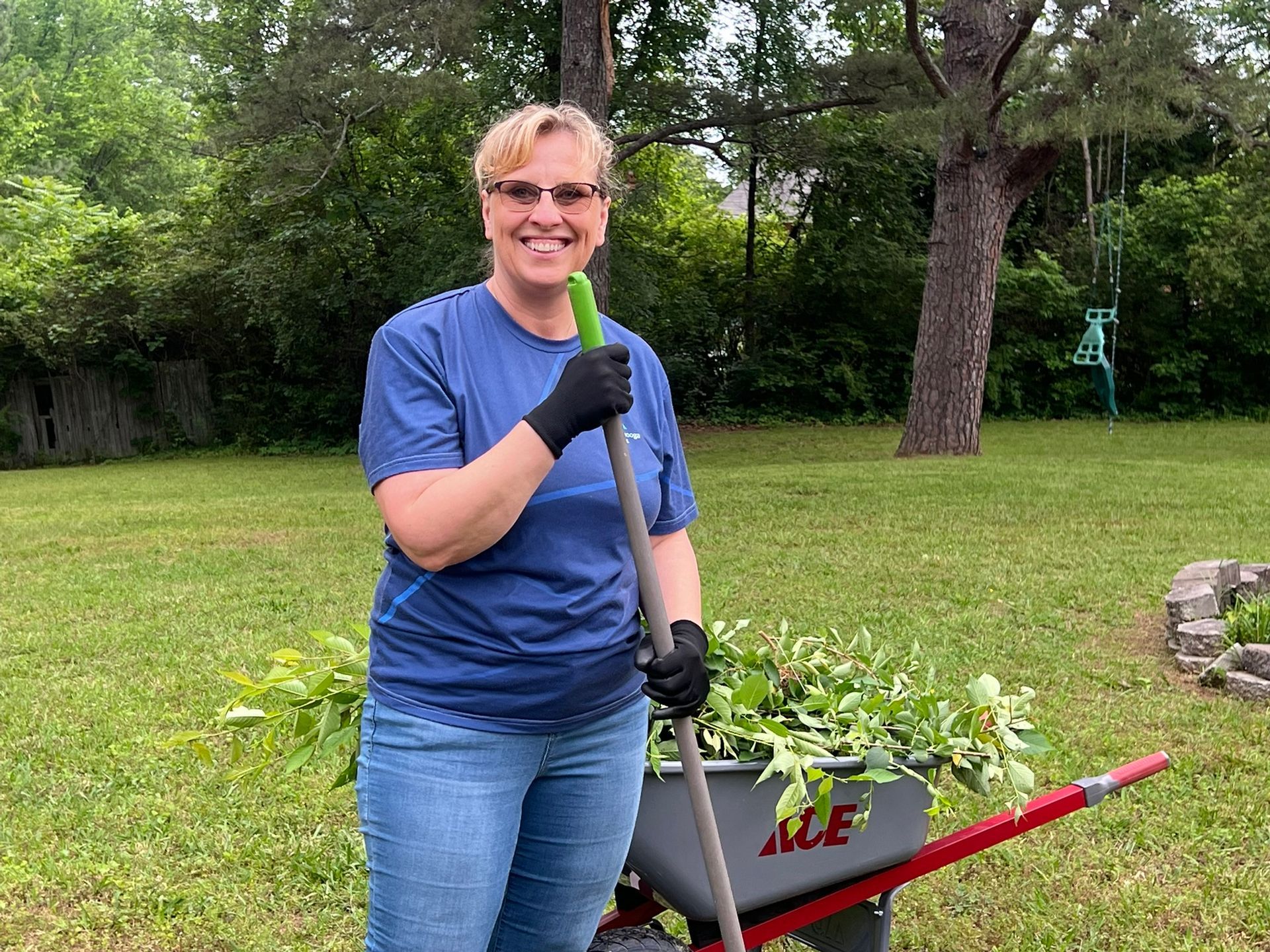 A female volunteer stands smiling, wearing gloves and holding a rake during a landscaping project.