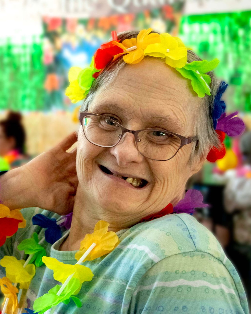 An older woman supported by Orange Grove Center smiles, wearing glasses and decorative floral leis for a luau celebration.