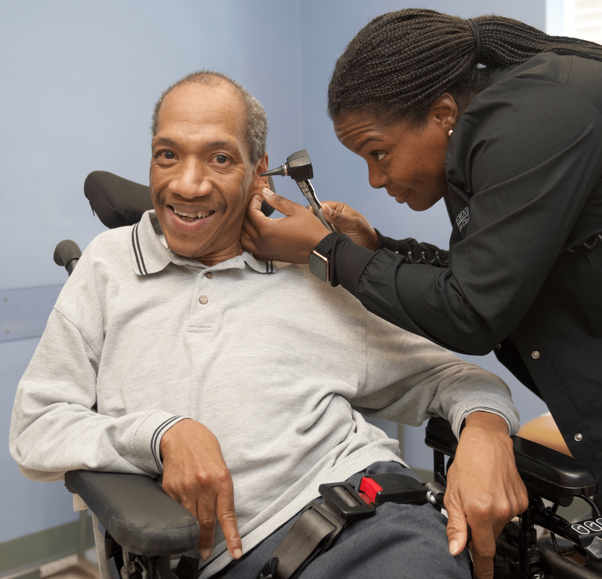 A man served by Orange Grove smiles while sitting in his wheelchair as his ear is inspected by a nurse.