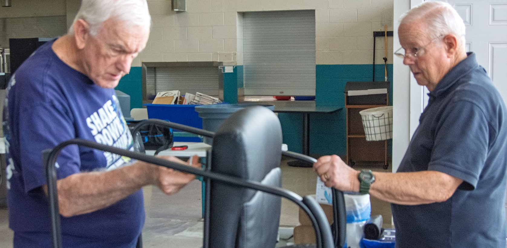 Two older male volunteers work together to assemble new chairs in the Orange Grove cafeteria.