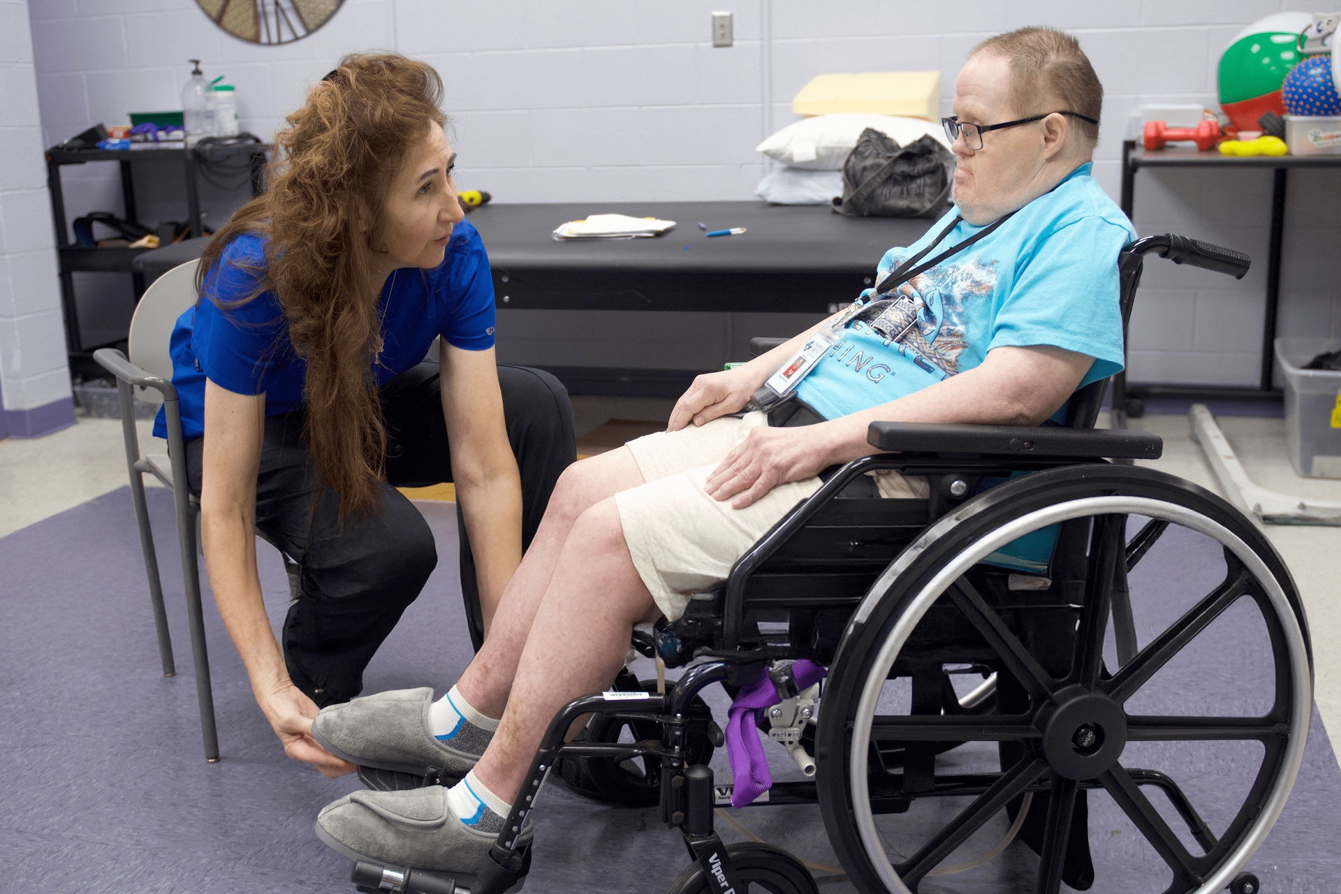 A man sits in a wheelchair, as a Clinical Services staff member assists him with his feet during physical therapy.