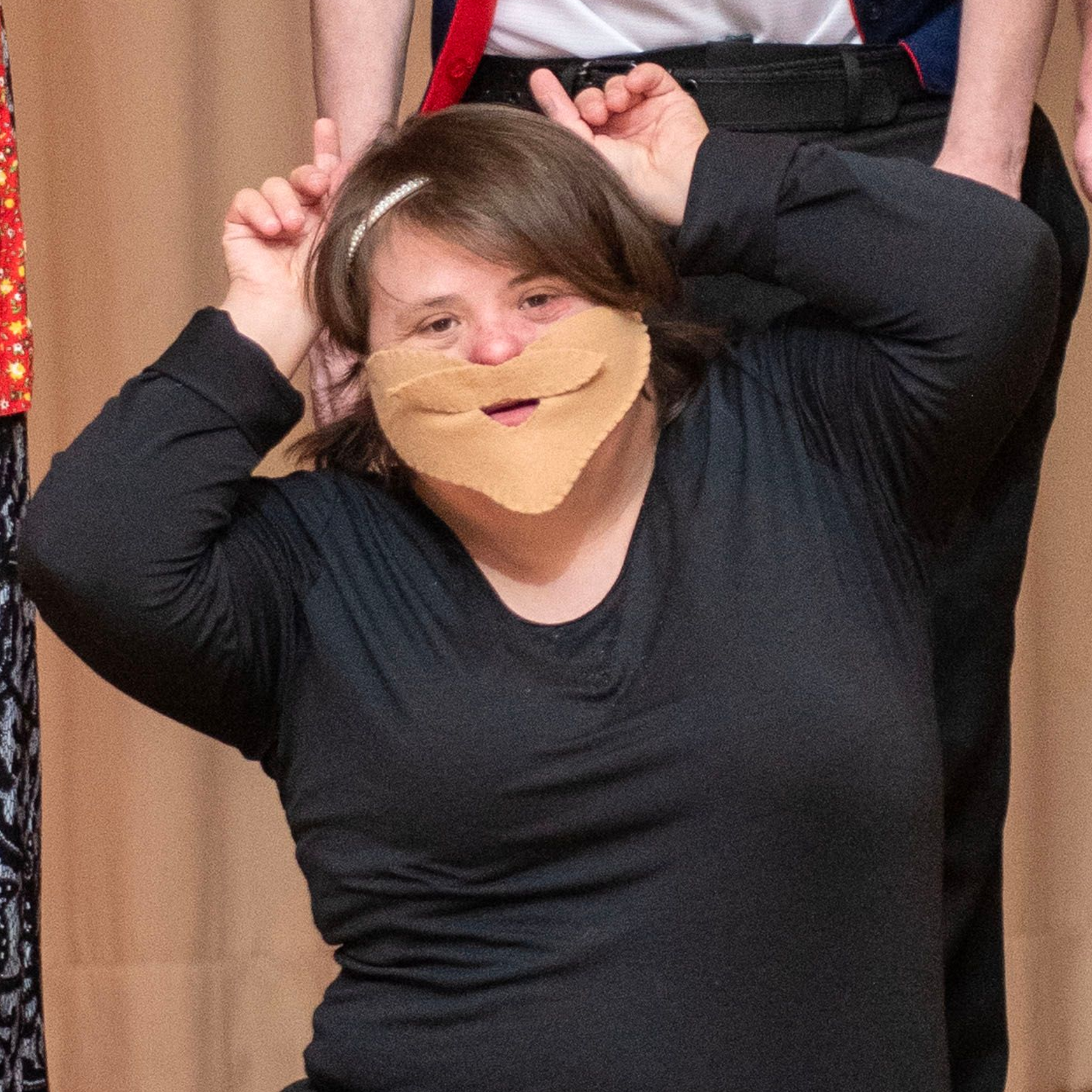 A woman wearing a bearded mask uses her hands to playfully mimic having pointed ears during a Summer Showcase performance.