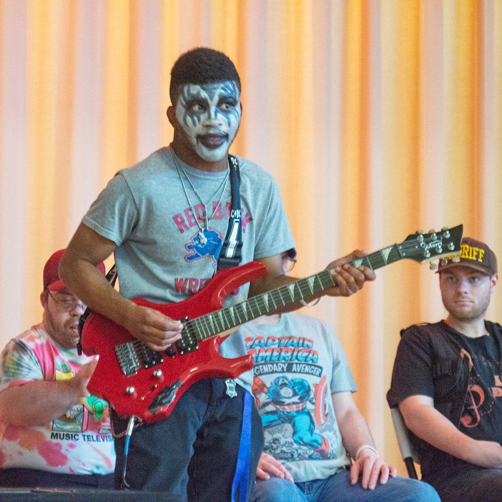 A man served by Orange Grove in KISS makeup, playing a red guitar during a concert.