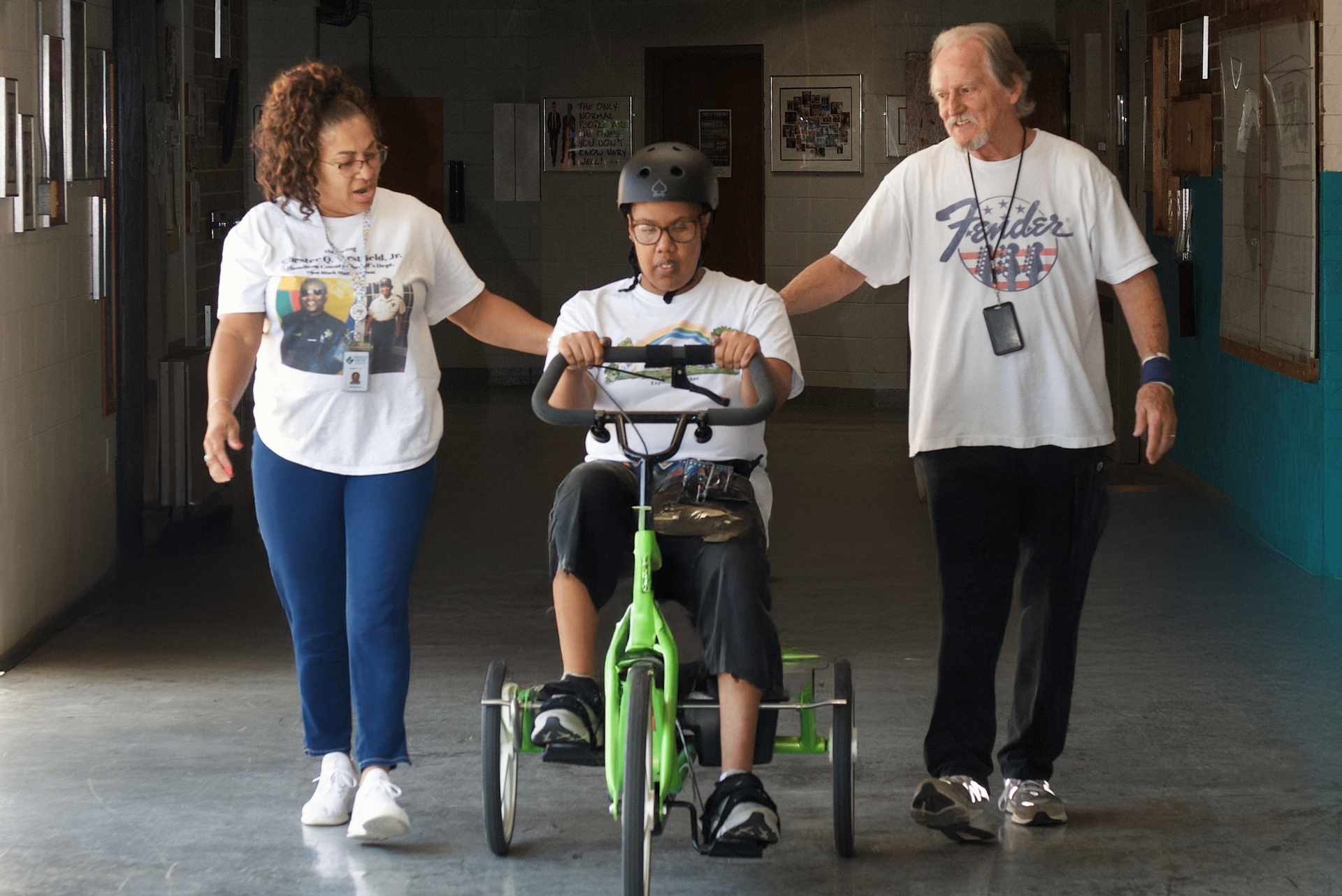 An individual served by Orange Grove pedals a therapy bike down a hallway, joined by clinical services staff.