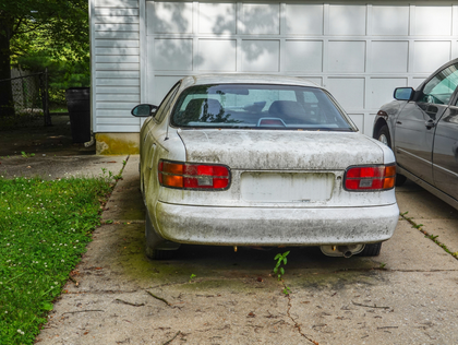 Dirty white car parked in a driveway beside a garage, with another car partly visible on the right.