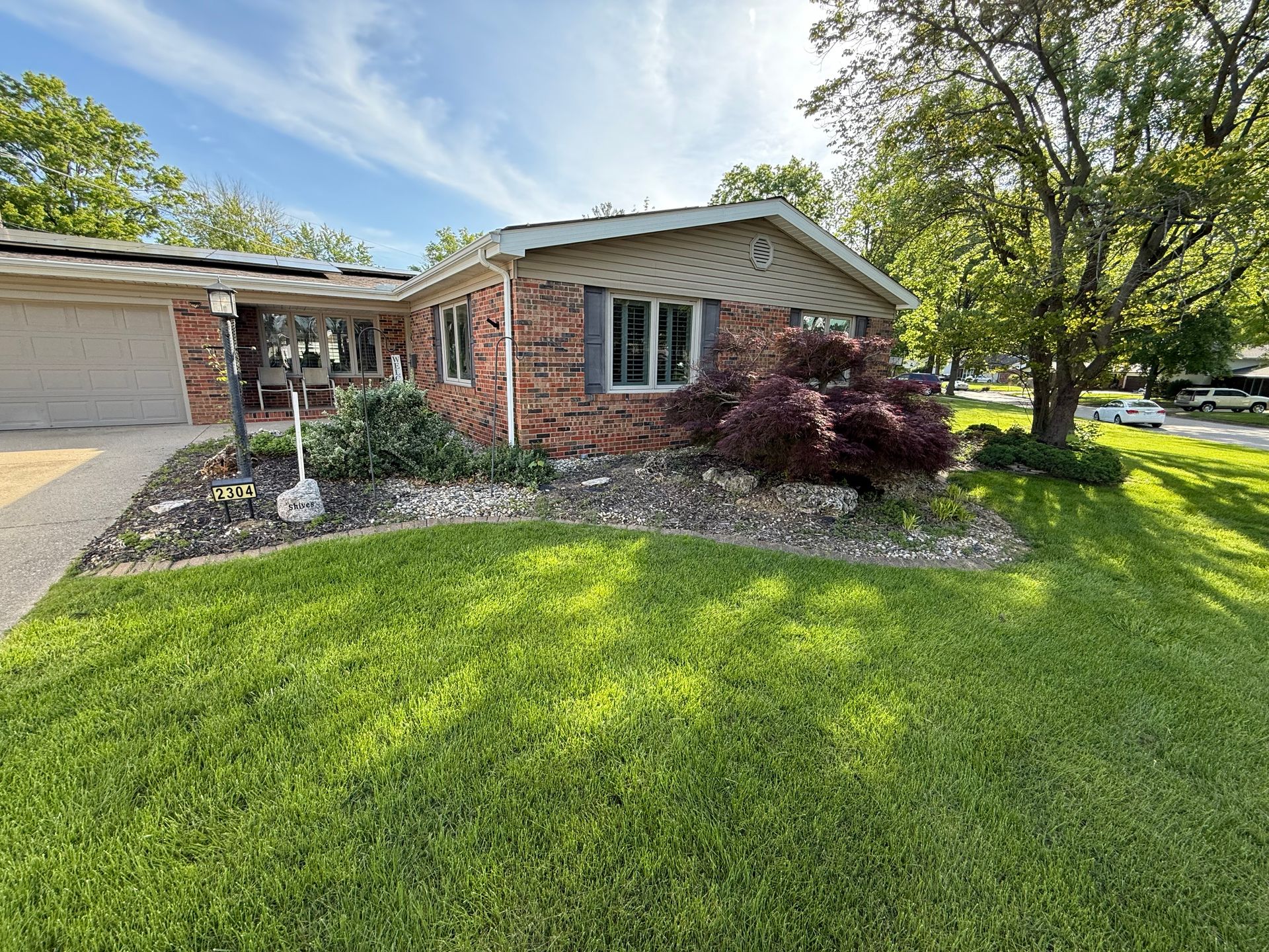 A single-story brick house with a front lawn, rock landscaping, a purple-leaf shrub, and a driveway on a sunny day.