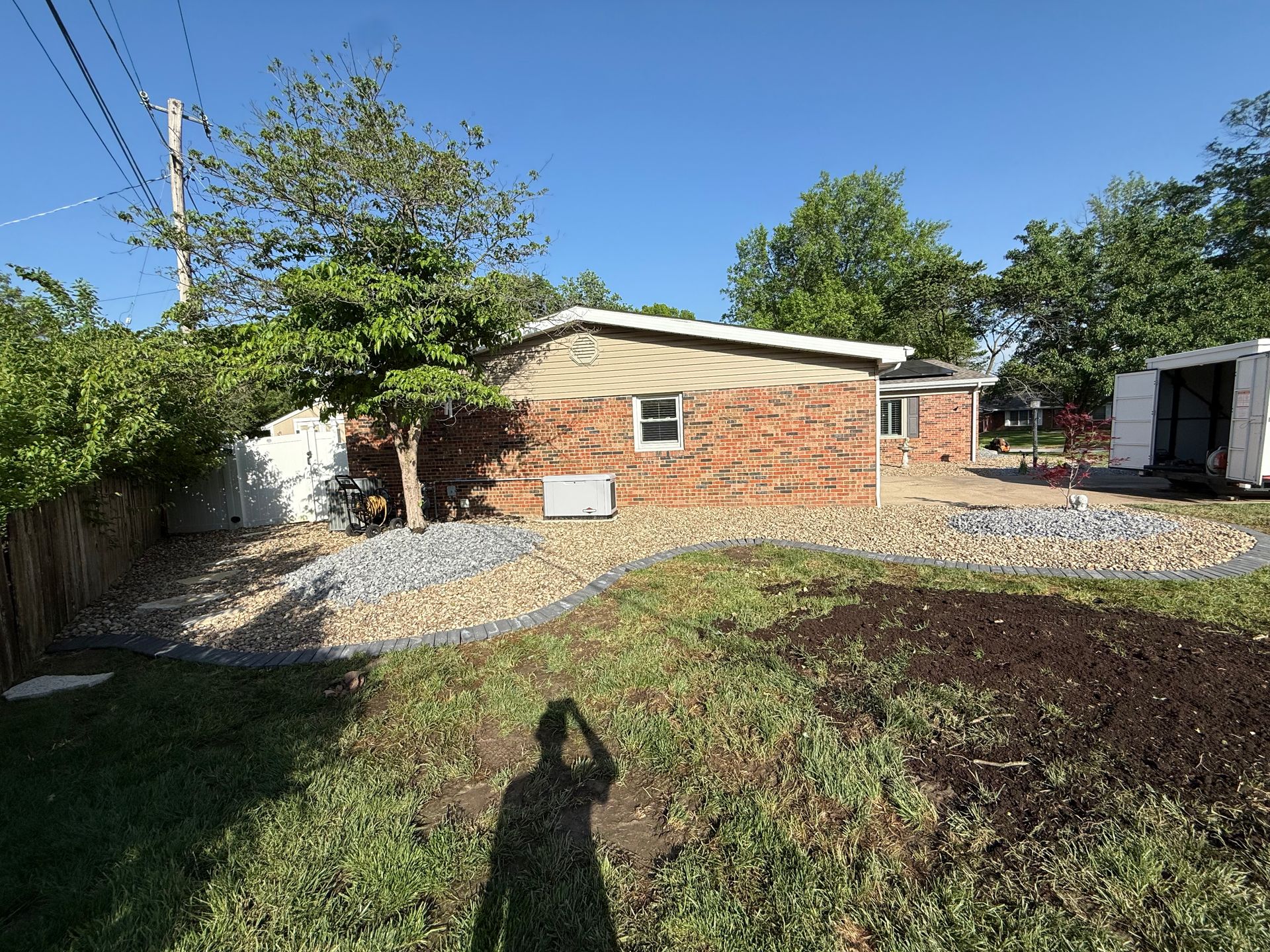 A backyard featuring a brick house with new landscaping, including curved stone beds, a patch of soil, and green grass.