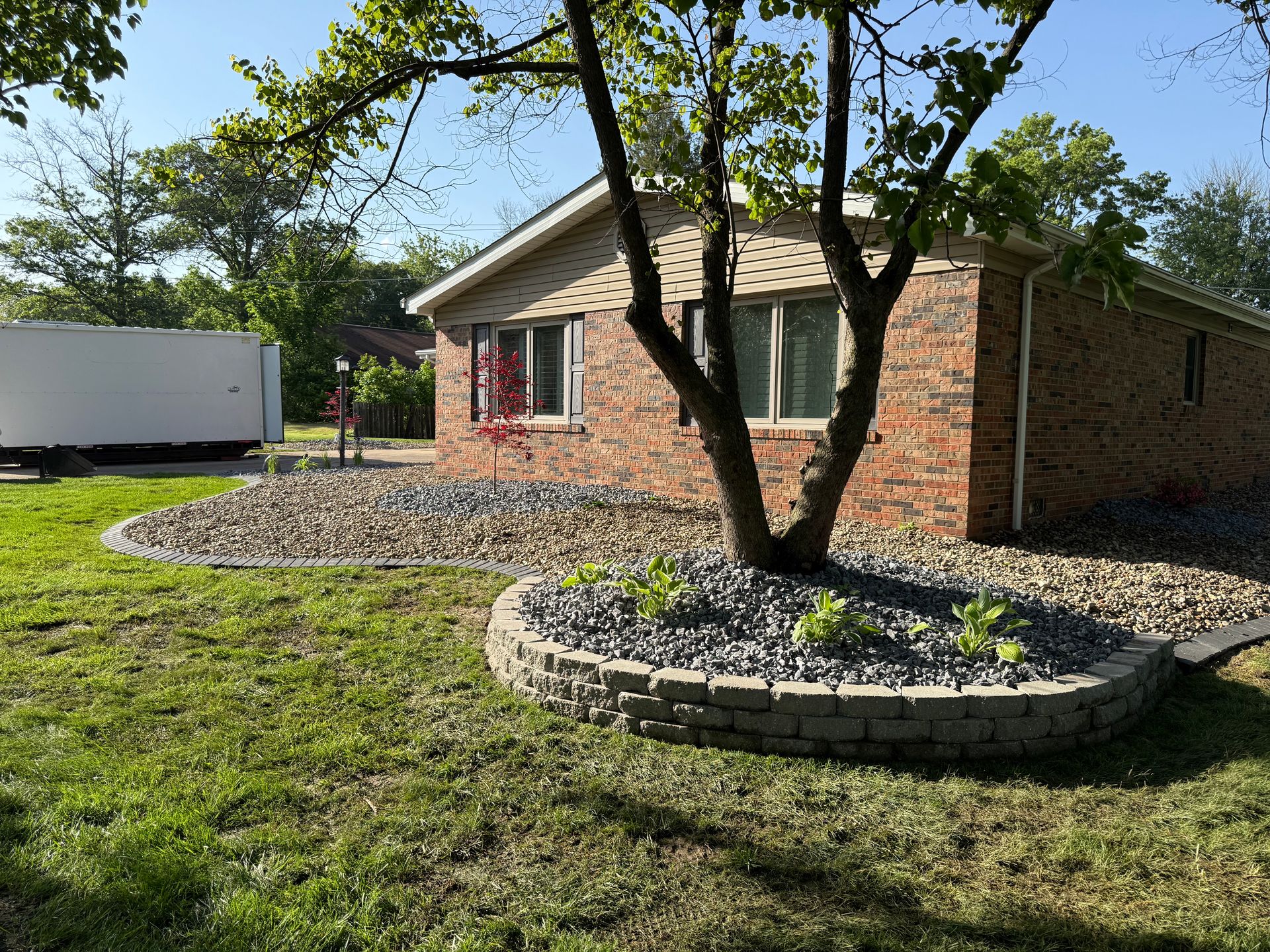 A brick house with a front yard featuring a raised stone garden bed around a tree and a gravel-covered landscape area.