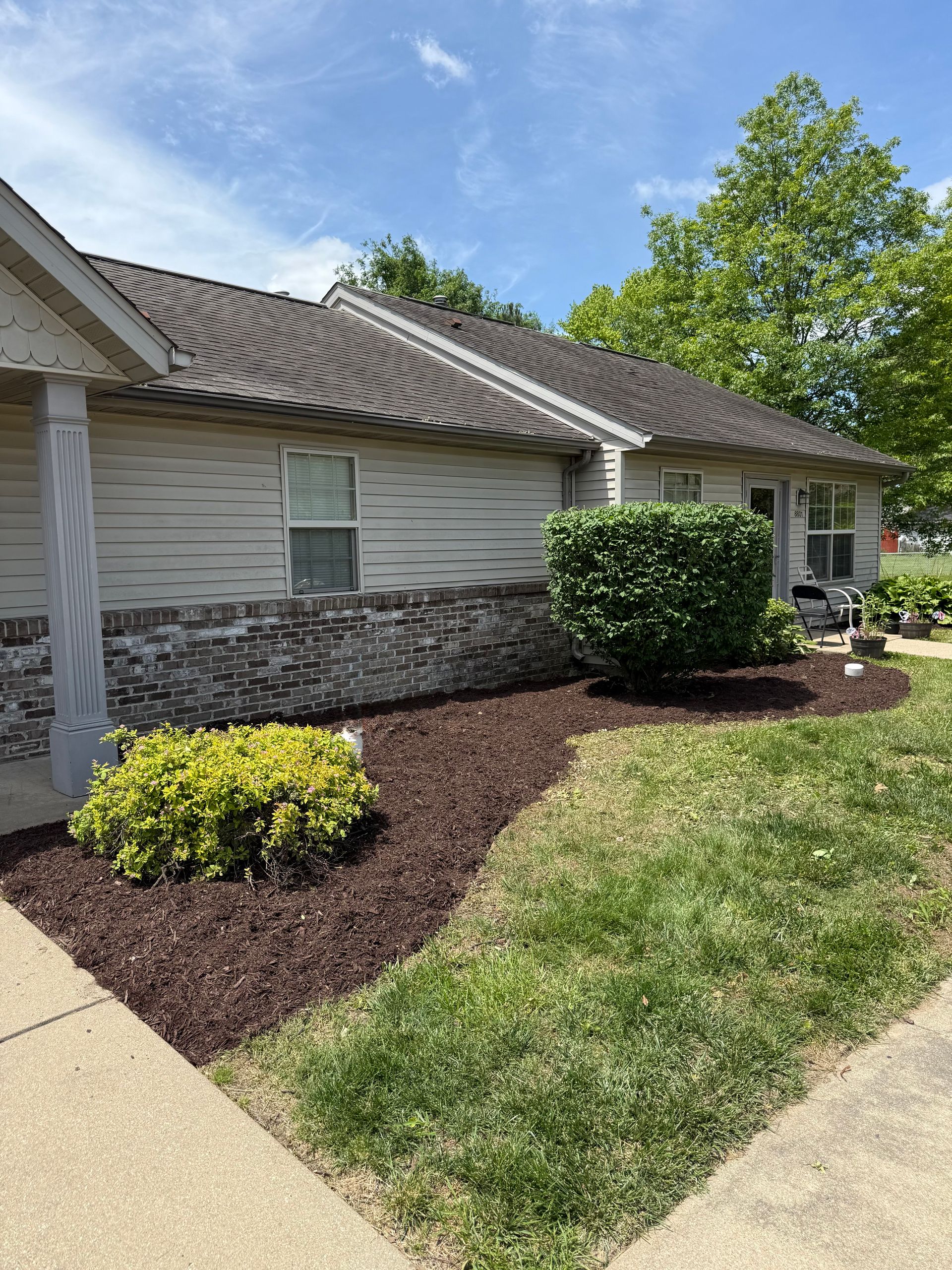 A single-story tan house with a brick base, brown shingled roof, dark mulch landscaping, and a manicured green lawn.
