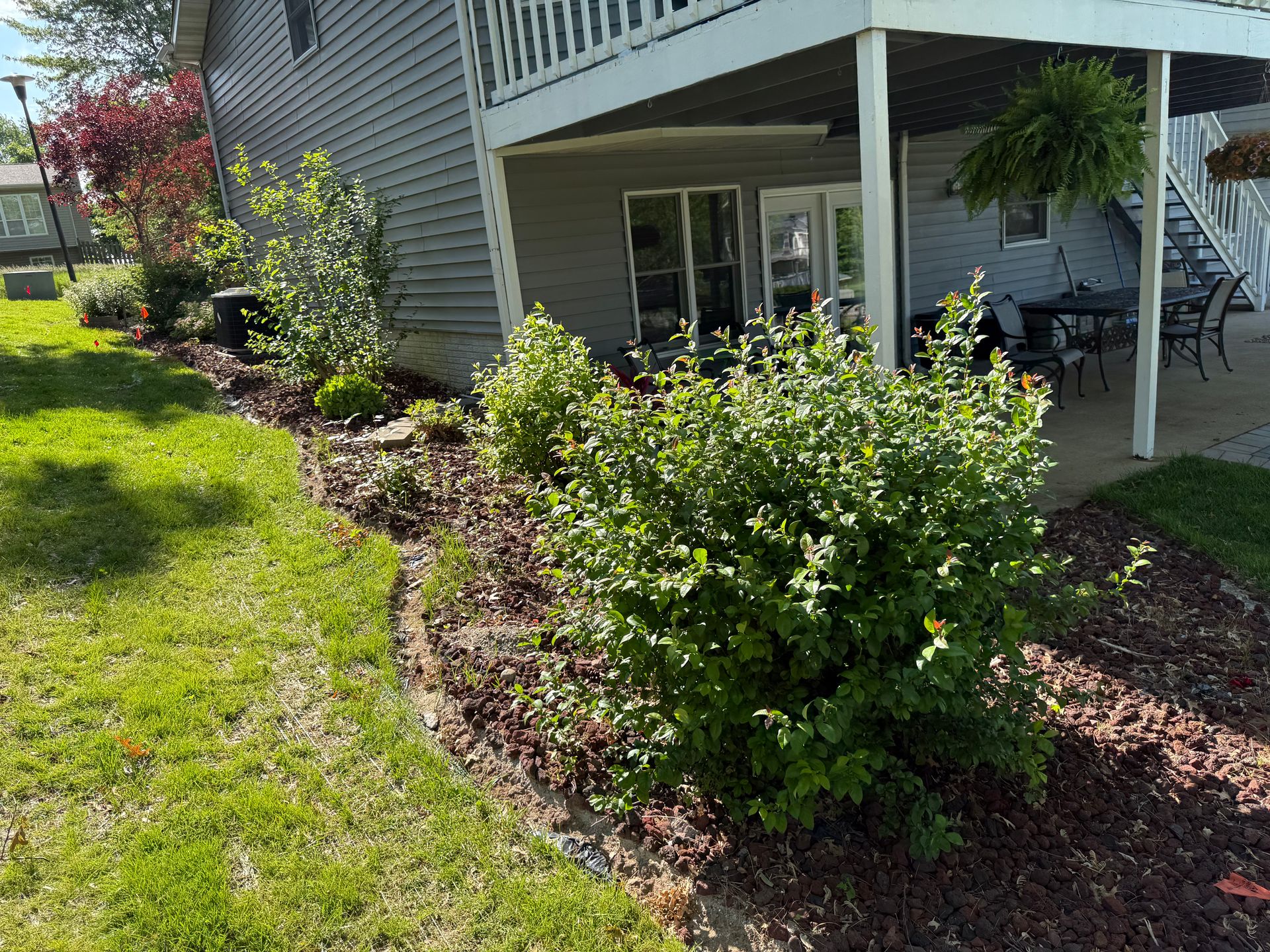 A side yard landscape with mulch beds, a large green shrub, and a wooden deck with patio furniture.