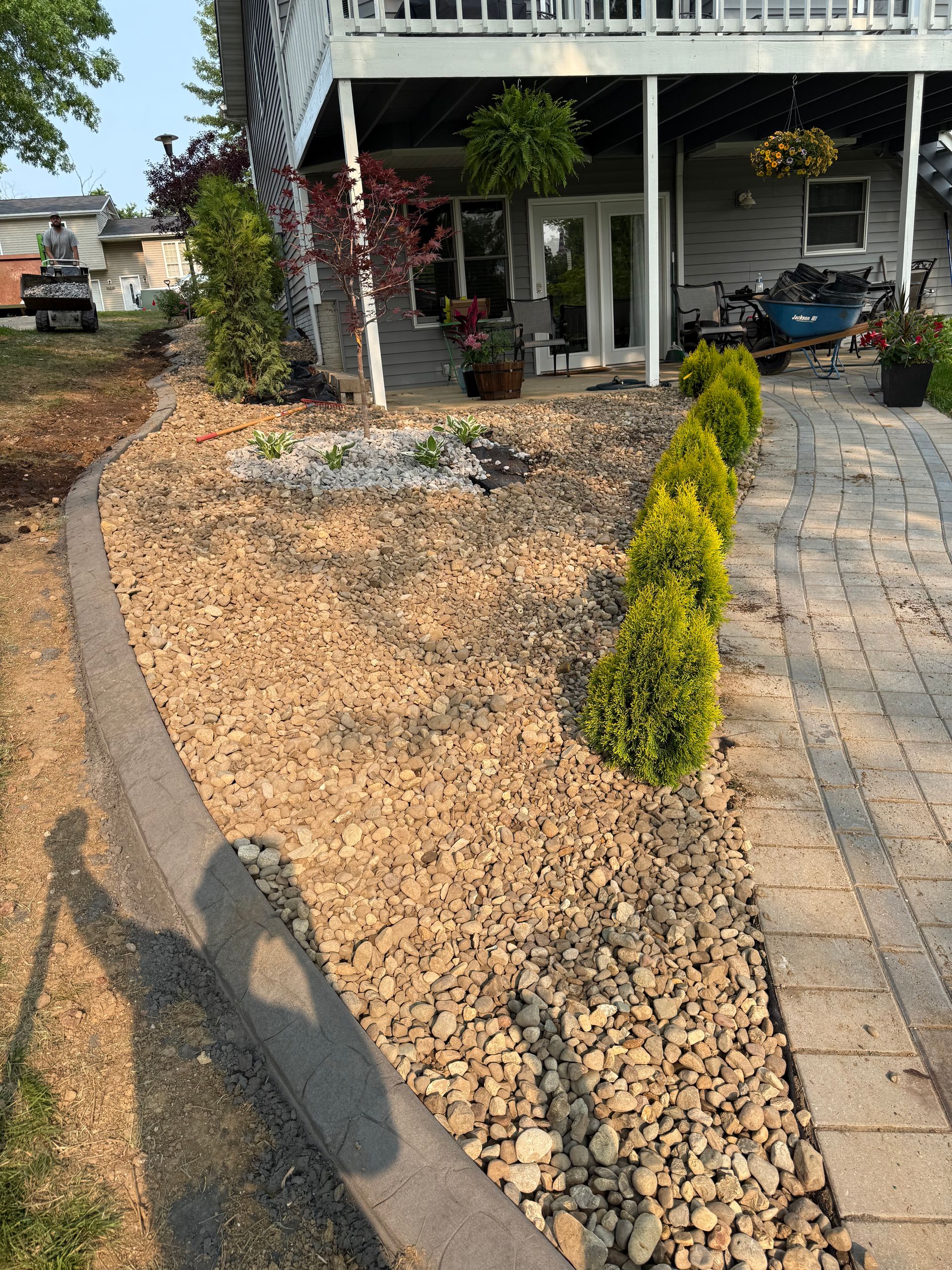 A stone walkway leads to a house patio, bordered by a rock garden with small shrubs and a concrete edge.