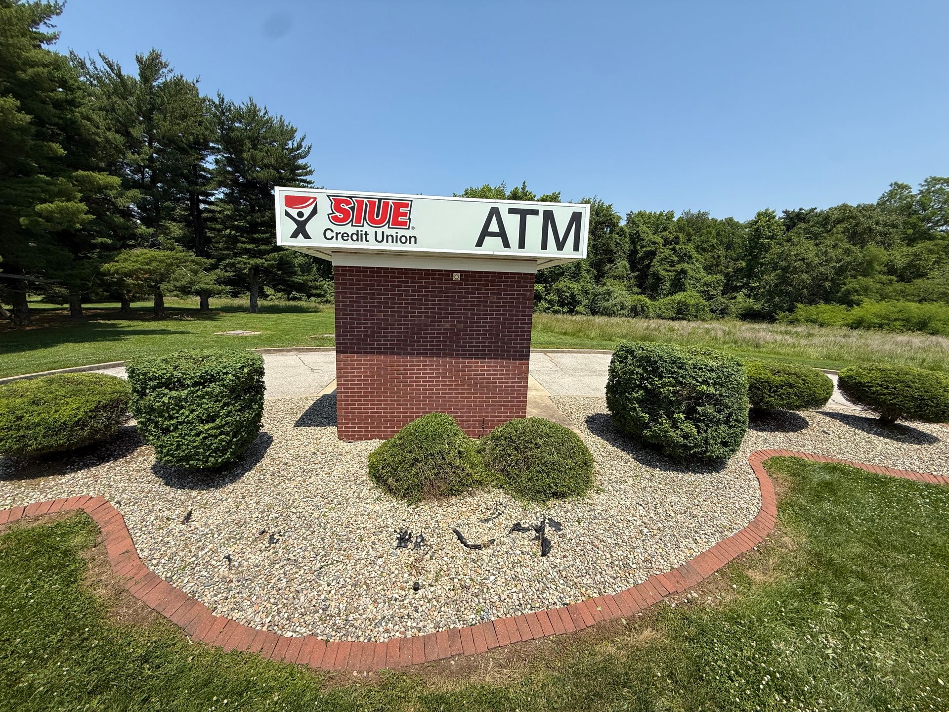 A S1NE Credit Union ATM sign on a red pedestal surrounded by gravel and manicured shrubs in a grassy, wooded setting.