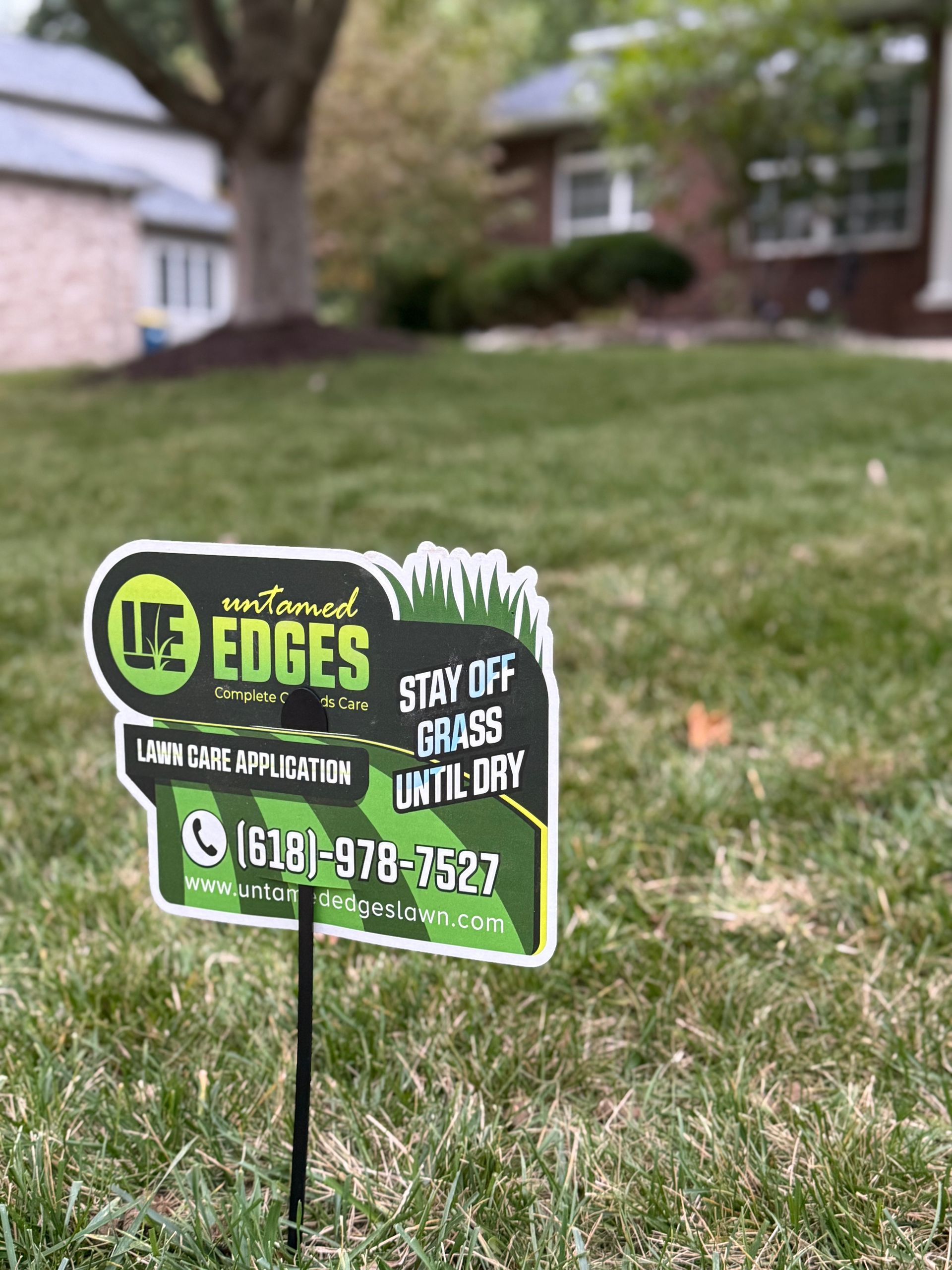 A green and black Untamed Edges lawn care sign placed in a grassy residential yard in front of a house.