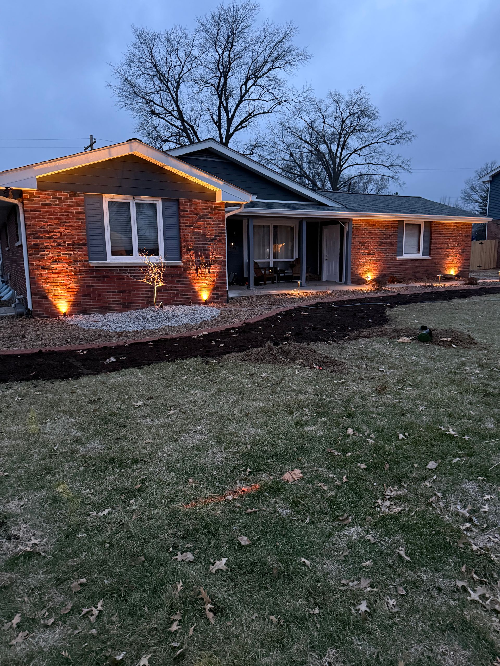 A single-story brick house at dusk, illuminated by warm exterior accent lights along the roofline and foundation.