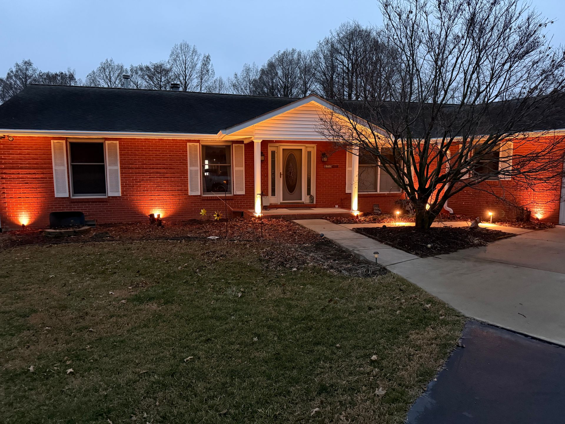 A single-story red brick house at dusk, illuminated by warm outdoor accent lighting along the exterior and front walkway.