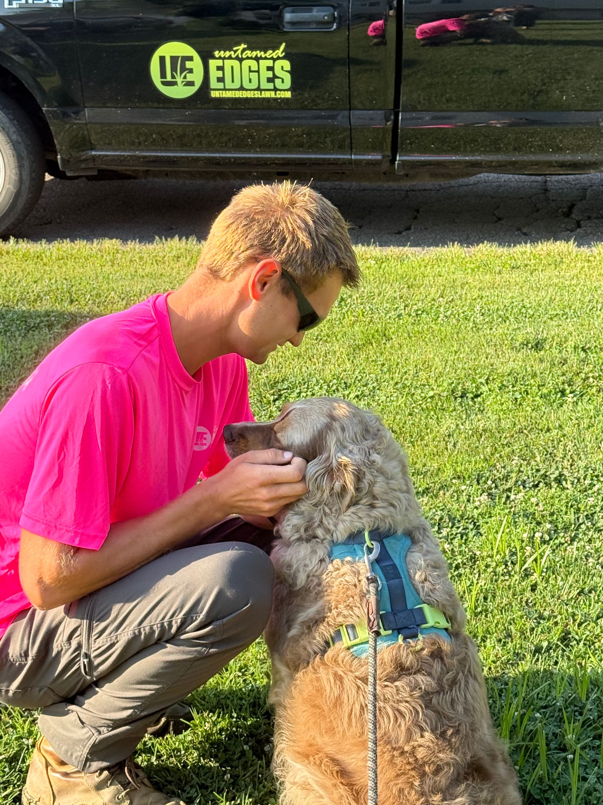 A person in a neon pink shirt kneels on the grass, gently petting a fluffy, tan-colored dog wearing a blue harness.