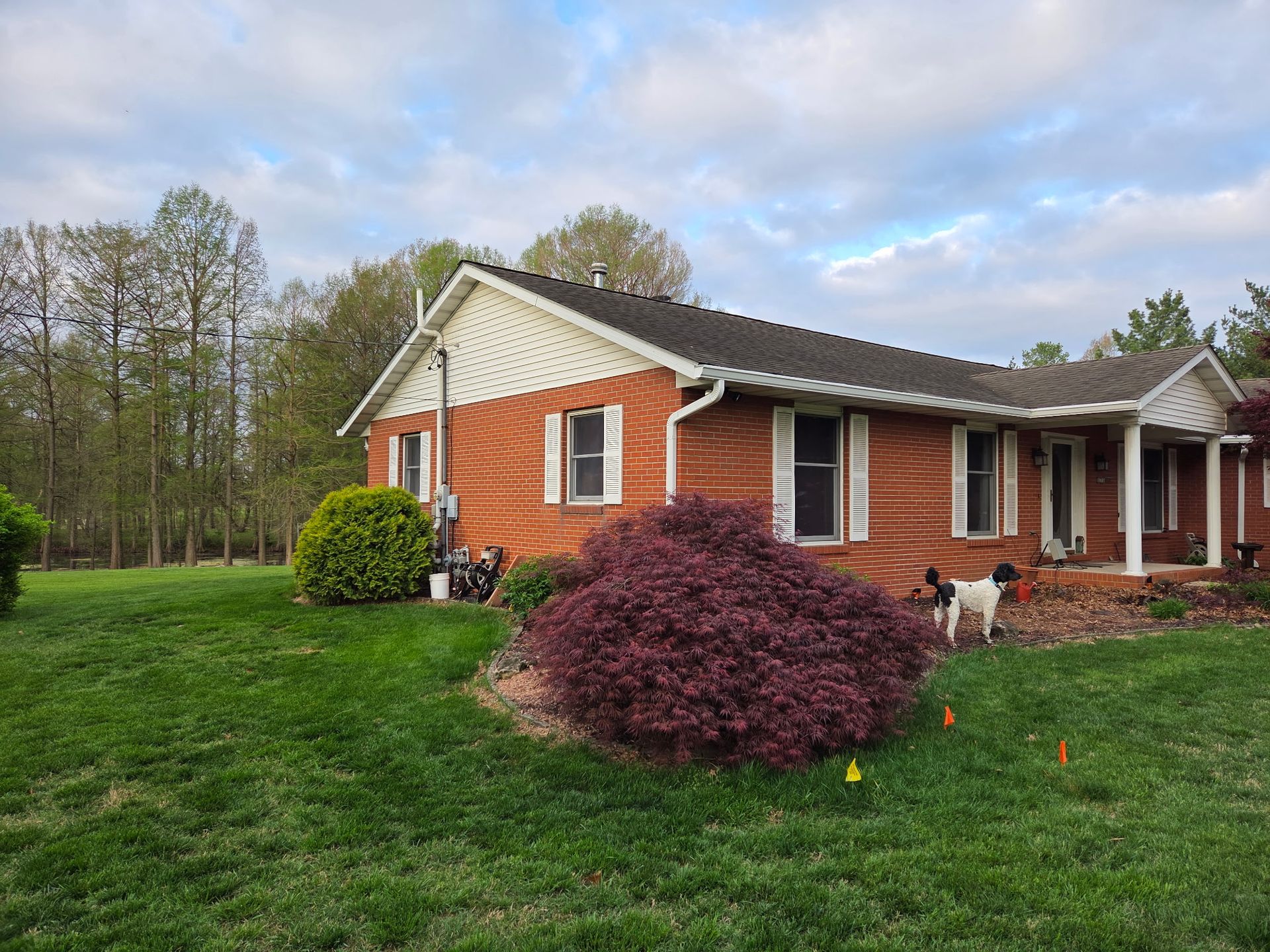 Brick house with white trim, a small porch, and a red shrub in a grassy yard under a cloudy sky