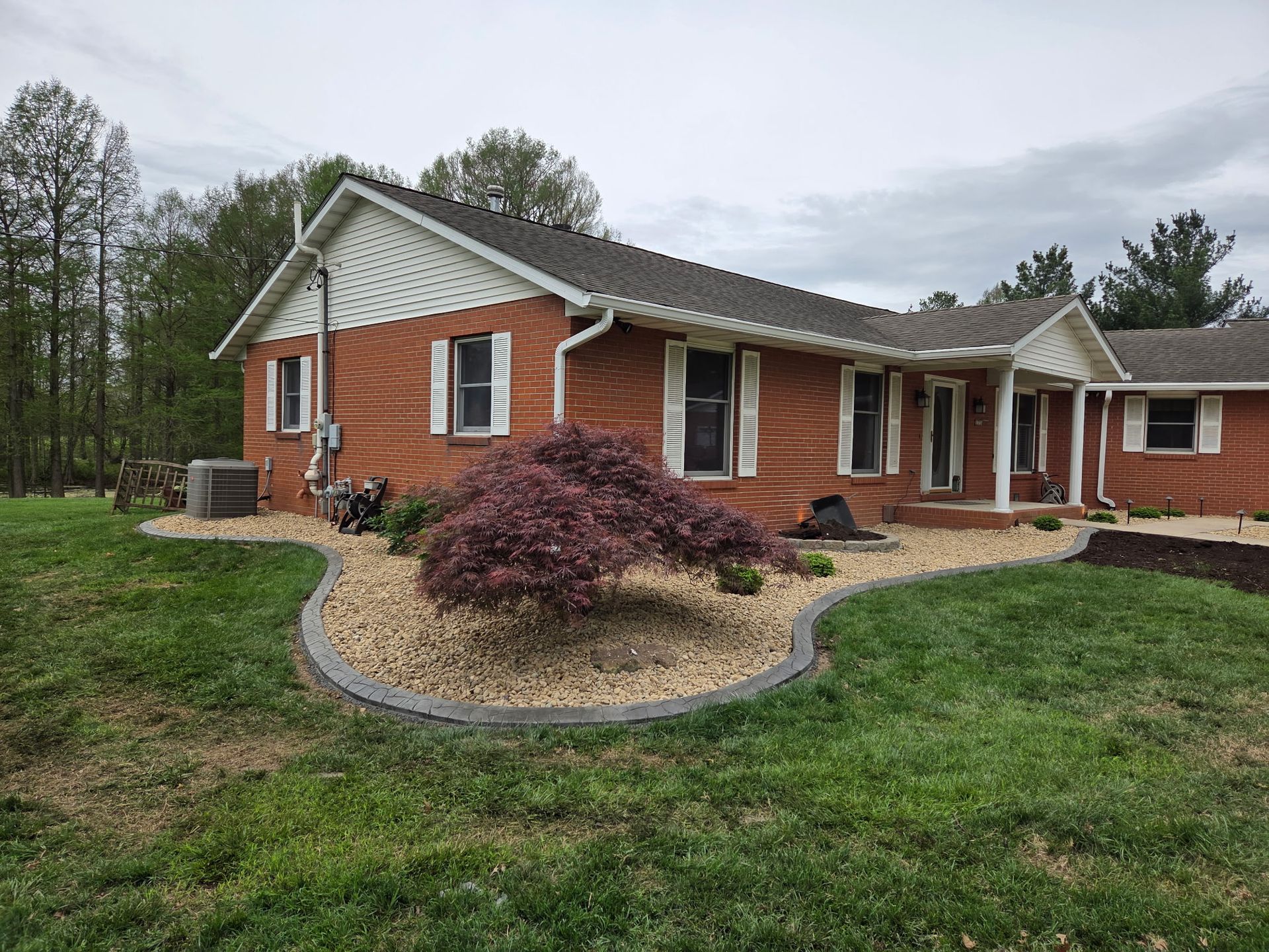 Brick ranch house with a curved stone bed, shrubs, and green lawn on a cloudy day