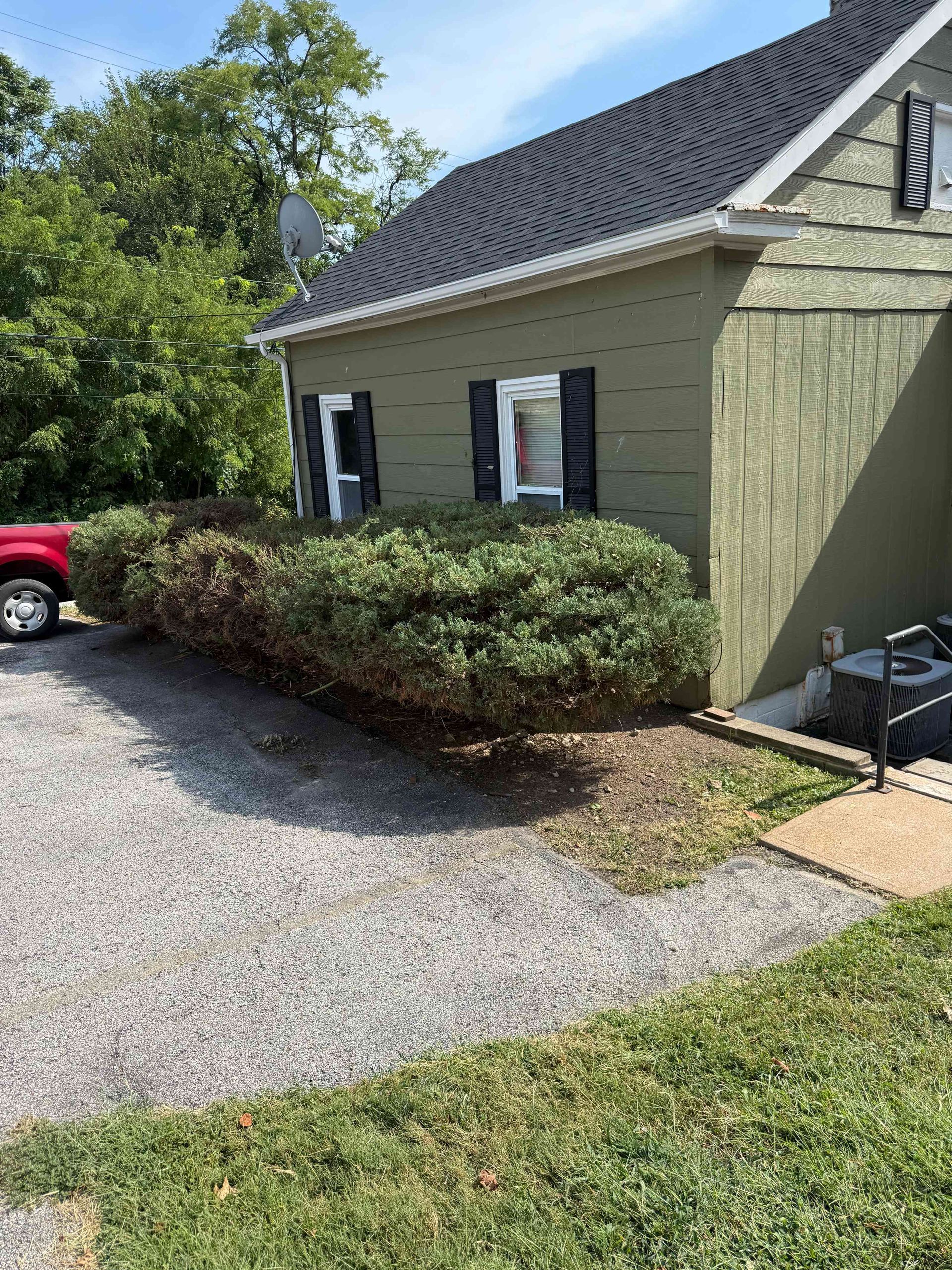 Small green house with white shutters, gravel driveway, bushes, and a red car partly visible on the left.