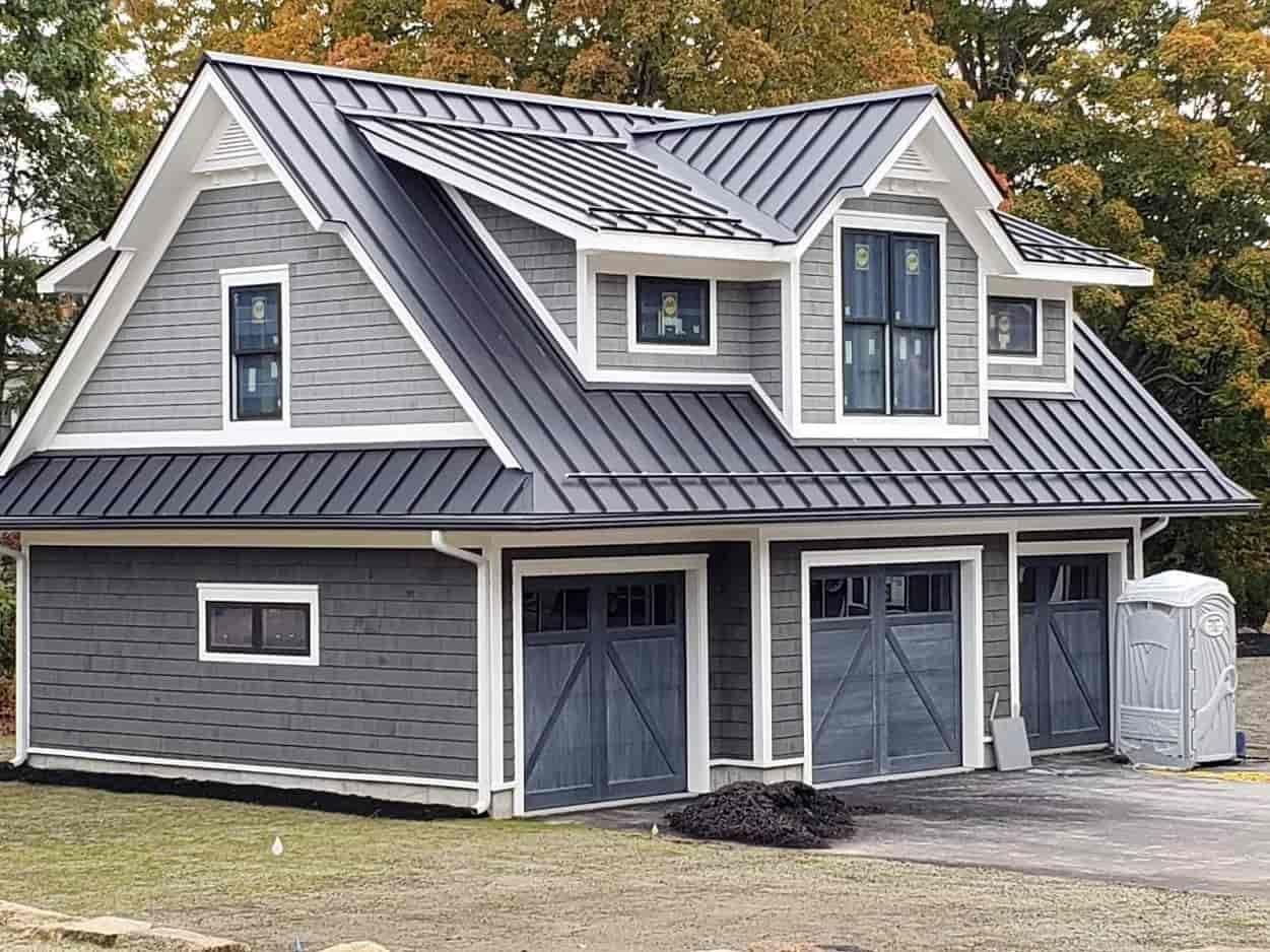 Gray two-story house with black metal roof, dormer windows, and front garage doors in a wooded yard