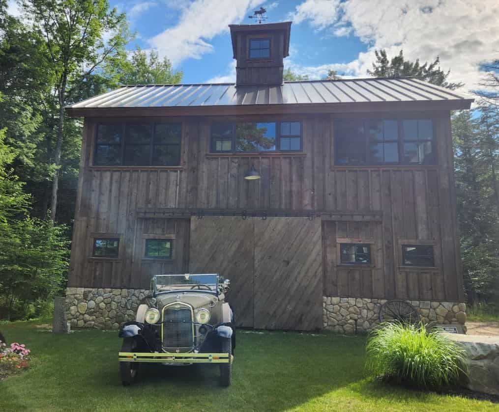 Rustic wooden barn with metal roof and cupola, set behind a vintage car on a green lawn under a partly cloudy sky
