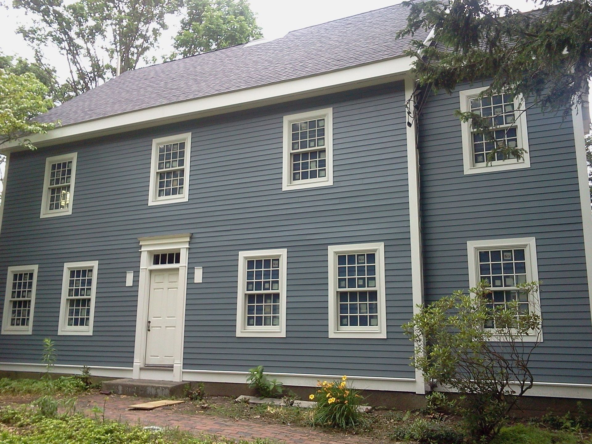 Blue-gray two-story house with white trim, many windows, and a front door surrounded by greenery