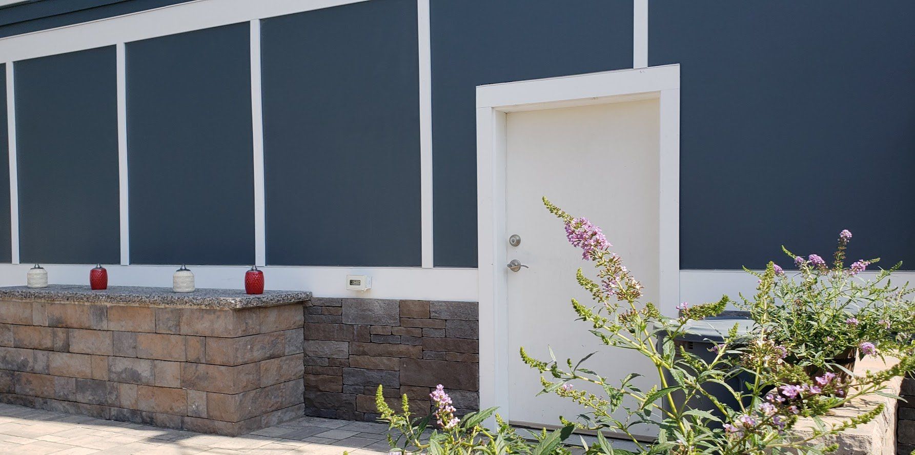 Modern building exterior with dark blue panels, stone wall, and white door by flowering shrubs