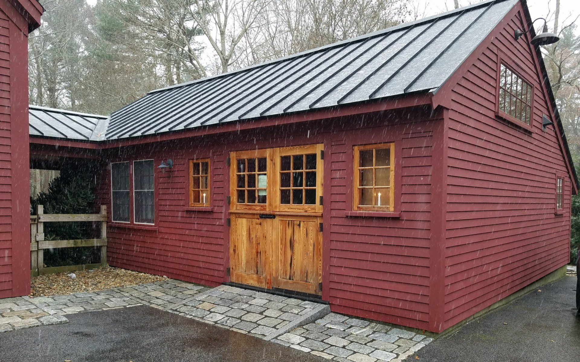 Red wooden cabin with double doors and a black metal roof, set beside a gravel driveway and trees.