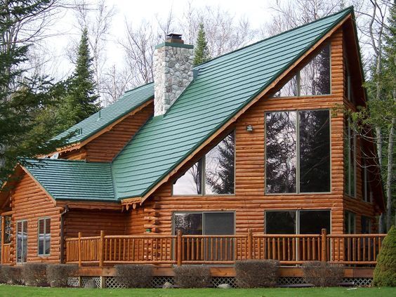 Wooden cabin with green metal roof and stone chimney in a wooded setting.