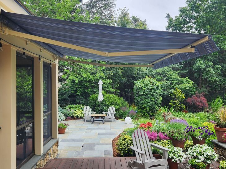 Patio with dark awning, stone walkway, white chairs, umbrella, and colorful flowers beside a house