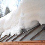 Snow piled on a brown, metal roof; a snowy forest is in the blurry background. Hot Edge | MasterCraft Roofing, Siding & Windows | Serving Eastern MA, NH & Selected Areas of VT