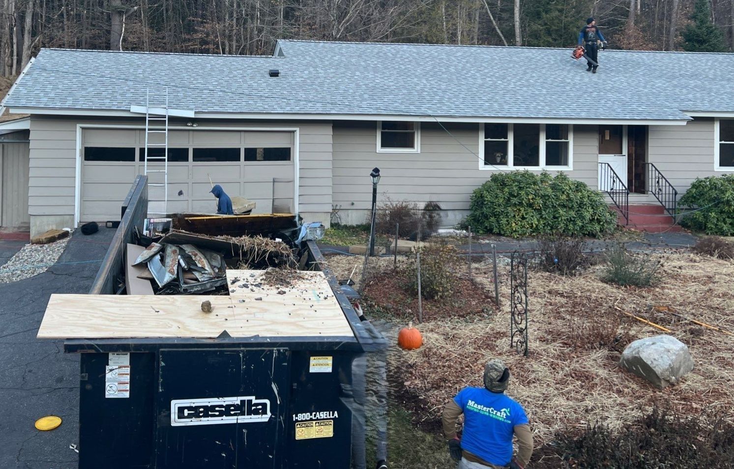 Single-story house with roof repair, a dumpster in front, and two workers on the roof and lawn