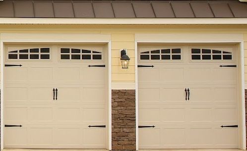 Two beige garage doors with small windows and a wall lantern in between