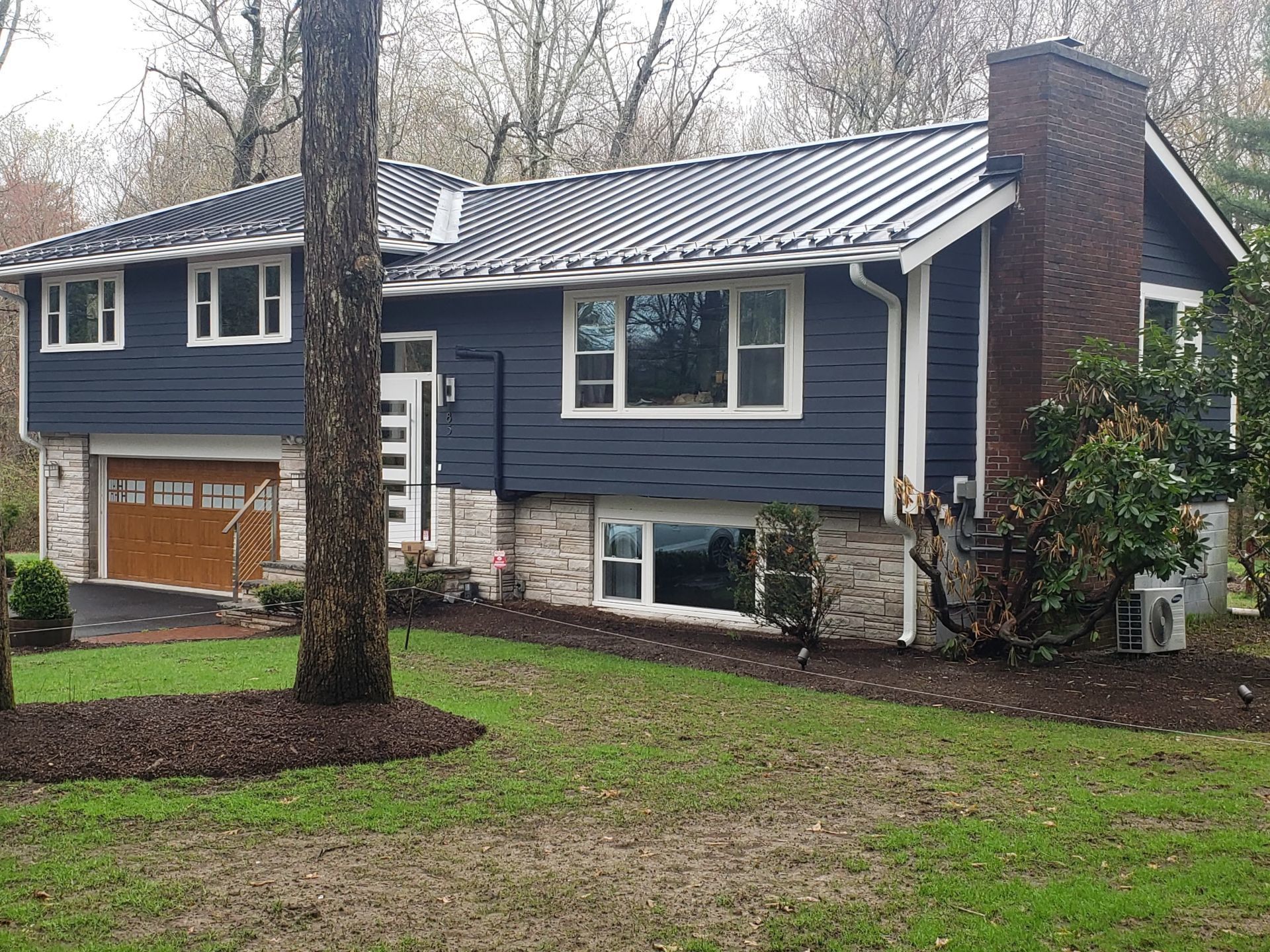 Blue split-level house with brown garage, white trim, and a brick chimney in a wooded yard