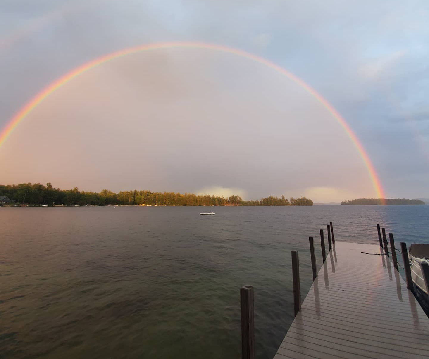 Rainbow over calm water and a dock at sunset, with a distant shoreline.