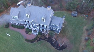 Aerial view of a large gray house with a front porch and landscaped yard surrounded by trees.