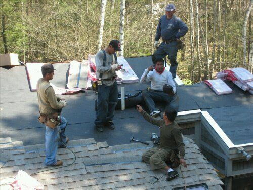 Workers repairing a shingled roof, with tools and materials spread across the rooftop.