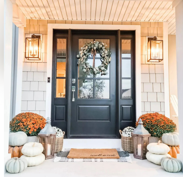 Black front door with fall wreath, lanterns, pumpkins, and orange mums on a porch.
