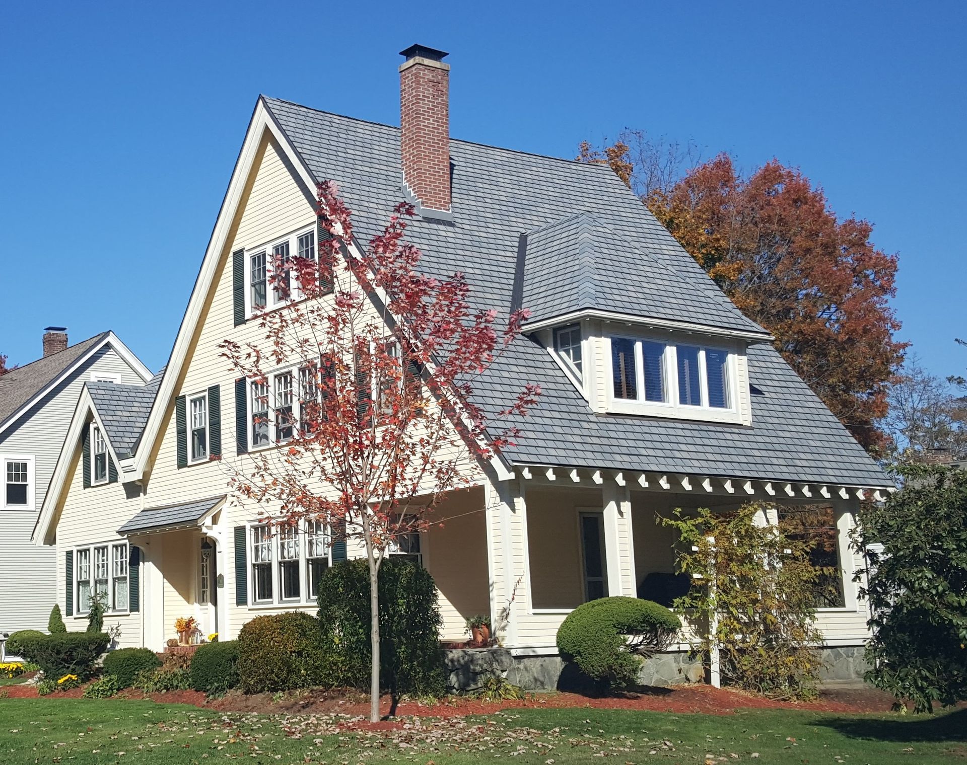Cream-colored house with gray roof, brick chimney, and autumn trees in front under a blue sky