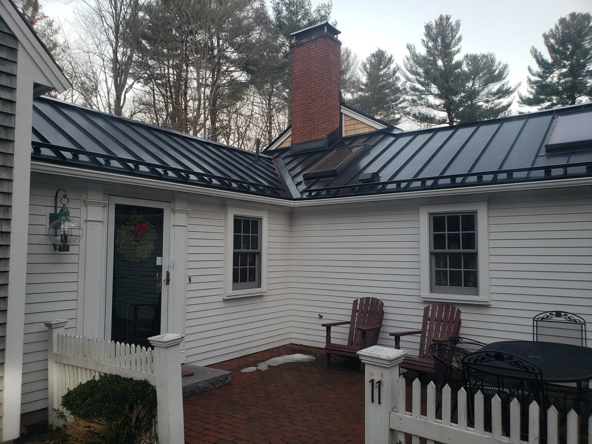 Brick courtyard with white house exterior, dark metal roof, chimney, and black railing on a wet patio