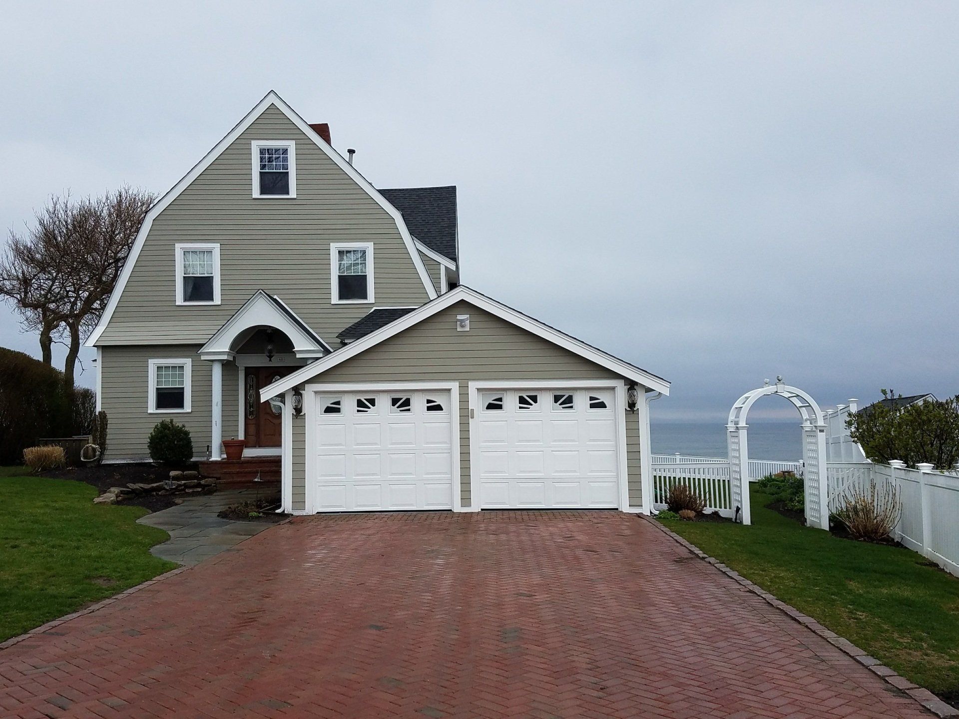 Gray house with white trim, double garage, brick driveway, and white garden arch by the water.