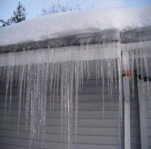 Icicles hanging from a snow-covered roof of a light-colored building in winter. Ice Dam | MasterCraft Roofing, Siding & Windows | Serving Eastern MA, NH & Selected Areas of VT