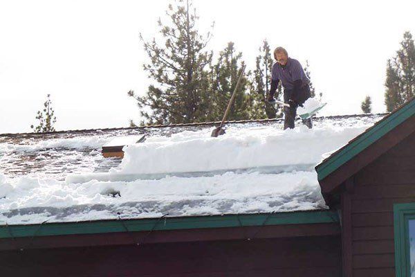 Person shoveling snow off a brown roof. Winter scene with trees in the background. Ice Dam | MasterCraft Roofing, Siding & Windows | Serving Eastern MA, NH & Selected Areas of VT