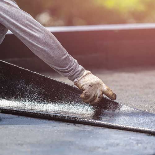 Gloved worker smoothing black roofing material on a rooftop in warm sunlight