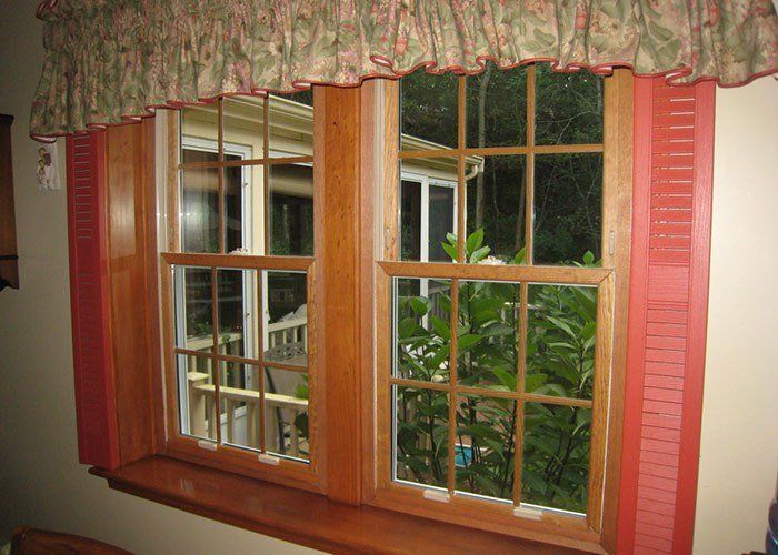Two wooden framed windows with red shutters and a floral valance. Trees visible outside. Energy Efficient | MasterCraft Roofing, Siding & Windows | Serving Eastern MA, NH & Selected Areas of VT