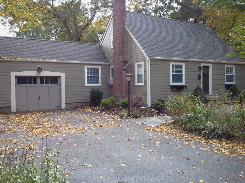 Gray house with attached garage, brick chimney, and leaf-covered driveway in a wooded yard
