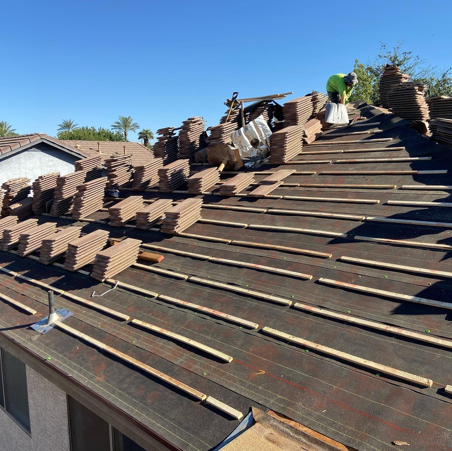 Roof being repaired with stacks of brown tiles on dark underlayment; worker in green shirt.