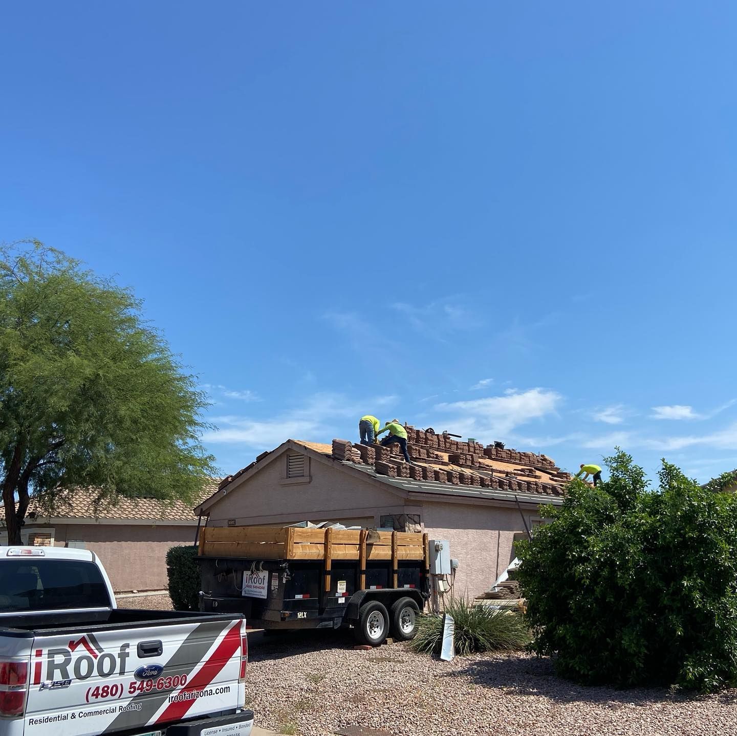 Roofers working on a house roof. Dump truck, white pickup, green bushes, blue sky.