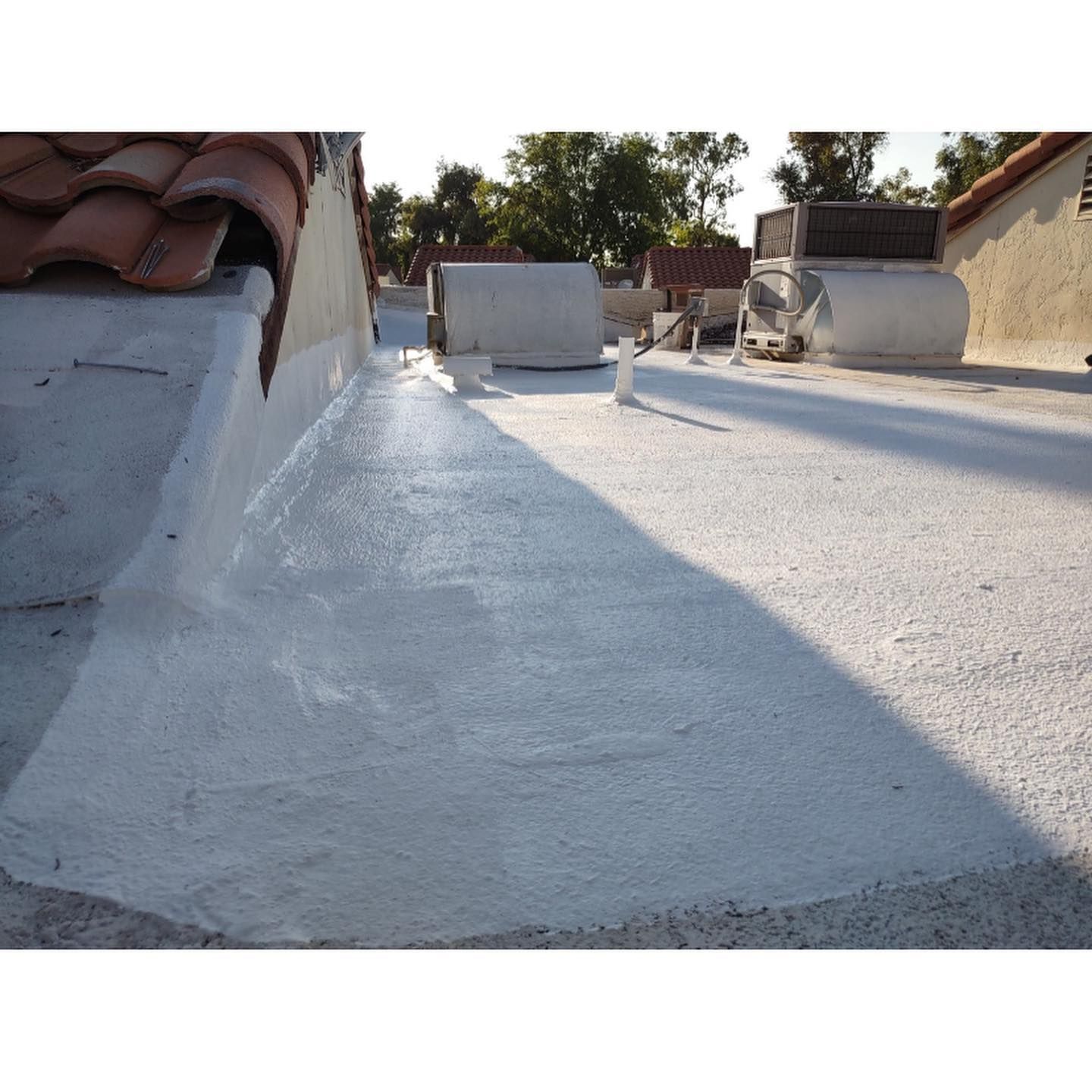 White-coated flat roof next to a tile roof, with some AC units in the background under a clear sky.