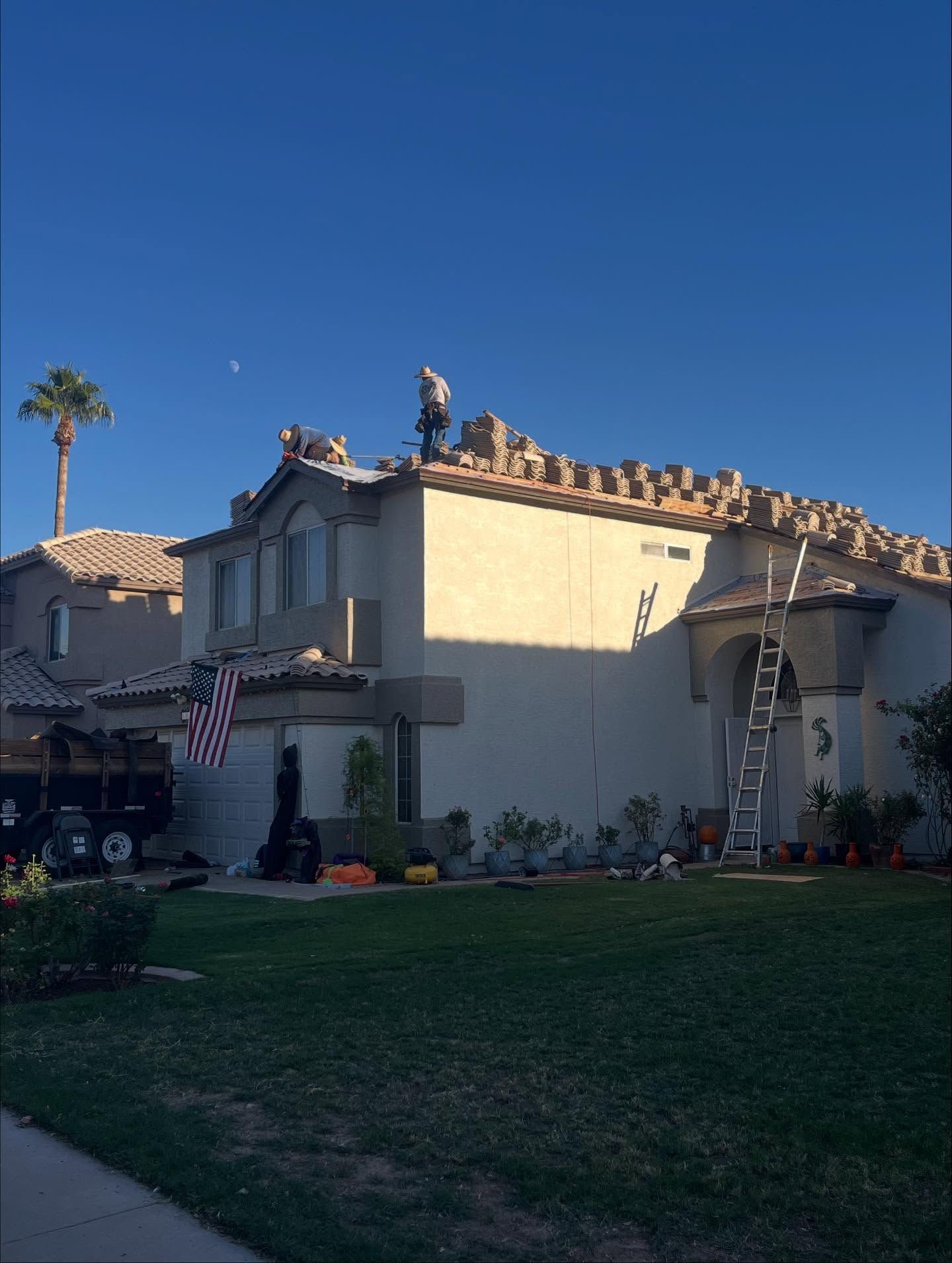 Two workers on a rooftop, replacing tiles.  Ladder up against the house; clear blue sky.